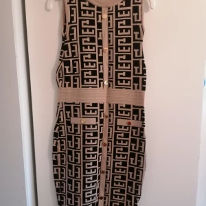 Beige och svart mönstrad ärmlös klänning - Snygg ärmlös klänning i beige och svart med grafiskt mönster och dekorativa guldknappar framtill. Klänningen har markerad midja och två fickor med knappdetaljer. Stickad känsla och rak passform, perfekt för dig som gillar statement-plagg.                                              Klänningen är stl S/M, men är sydd vid underarmarna för att passa xs. 