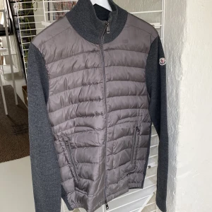 Grå Moncler cardigan jacka med dun - Snygg grå cardigan jacka från Moncler med quiltad framsida i dun och stickade ärmar och rygg. Jackan har hög krage, dragkedja framtill och två fickor med dragkedja. Storlek L. Jättebra sparsamt skick. Säljes pga för liten nu.