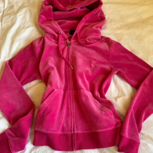 Juicy Couture rosa velour hoodie - Juicy Couture hoodie i starkt rosa velour med dragkedja, huva och snörning. Klassisk JC-logga broderad på bröstet och fickor framtill. 