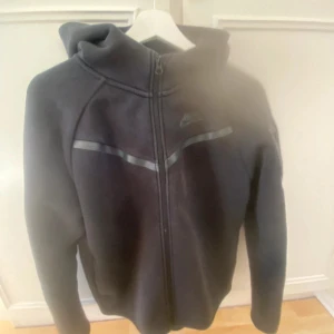 Svart Nike hoodie med dragkedja - Svart hoodie från Nike med hel dragkedja framtill och huva. Tröjan har en diskret Nike-logga på bröstet och en reflekterande detalj över bröstet. Perfekt för en sportig och avslappnad stil.