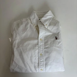 Vit skjorta från Ralph Lauren - Klassisk vit skjorta från Ralph Lauren med broderad logga på bröstet. Skjortan har lång ärm, knappar framtill och en snygg custom fit-passform. Tillverkad i mjuk bomull som känns skön mot huden. Perfekt för dig som gillar stilrena och tidlösa plagg.