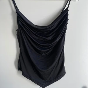 Svart draperad linnetopp med smala band - Säljer en svart linnetopp med smala, justerbara axelband, stretch och snygg drapering framtill. Toppen har en lätt glansig finish och spetsig nederkant, vilket ger en trendig look. Perfekt att styla med jeans eller kjol för en cool outfit. 