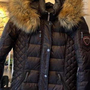 Svart lång dunjacka Rockandblue - .Snygg figursydd jacka i dun från ROCKANDBLUE. Stl 36 Säljes pga av att jag växt ur den.  Knappt använd! Köpte den för 3199kr  Jackan har fina detaljer och stängs med dragkedja och tryckknappar framtill.  - Tre fodrade fickor framtill samt en liten ficka på ena armen med logotyp 