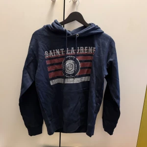 Blå hoodie Saint Laurent - Blå hoodie från Saint Laurene med tryck i rött, vitt och blått framtill. Tröjan har huva med snörning och en stor magficka. Perfekt för dig som gillar en sportig och avslappnad stil.