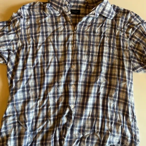 Rutig kortärmad skjorta från Randolph - Kortärmad skjorta från Randolph i blått och vitt rutigt mönster. Skjortan har klassisk krage, knäppning framtill och en bröstficka. Tillverkad i mjuk bomull, perfekt för en avslappnad stil.