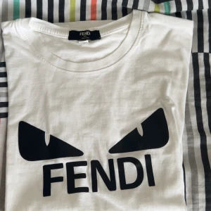 Vit Fendi t-shirt med svart tryck - Vit t-shirt från Fendi med ikoniskt svart monsterögon-tryck och FENDI-logga framtill. Klassisk rund halsringning och mjukt bomullsmaterial. Perfekt statement-plagg för dig som gillar exklusiva märken och cool design.