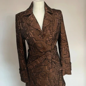 Brun blommig kappa från Mimar Collection - Snygg brun kappa från Mimar Collection med blommigt mönster i svart och beige. Kappan har klassisk krage, knäppning framtill och bälte i midjan. Långärmad och fodrad för extra komfort. Perfekt för dig som vill sticka ut med en unik och trendig look.