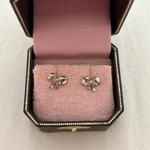 Juicy Couture örhängen med rosett - Snygga silverfärgade örhängen från Juicy Couture i form av små rosetter med en glittrande sten i mitten. Perfekt accessoar för att ge din look lite extra bling och söt vibe. Kommer i original ask med lyxig rosa insida och gulddetaljer.