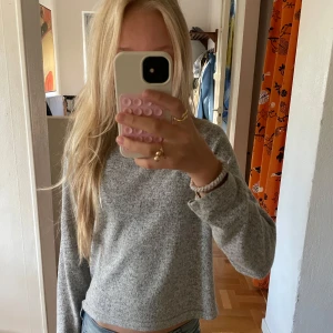 Grå långärmad stickad tröja - Mysig grå stickad tröja med lång ärm och avslappnad passform. Perfekt för kyliga dagar och enkel att matcha med jeans eller kjol. Tröjan har en klassisk rund hals och är lite croppad i modellen.