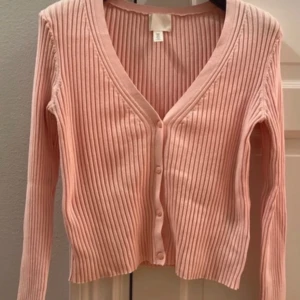 Rosa ribbad kofta med v-ringning - Snygg ljusrosa kofta från H&M med ribbad struktur och v-ringad hals. Modellen är croppad och har långa ärmar samt knappar framtill. Perfekt att styla med jeans eller kjol för en chill look. Är Strlk M men sitter som S