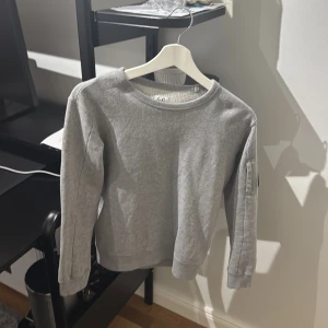 Grå sweatshirt från C.P. Company - Säljer en stilren grå sweatshirt från C.P. Company med klassisk rund hals och ribbade muddar. Tröjan har en ikonisk svart linsdetalj på ärmen och är tillverkad i mjuk bomull för maximal komfort. Perfekt för dig som gillar streetwear och snygga basplagg. Storlek 10 passar 165/49 kg