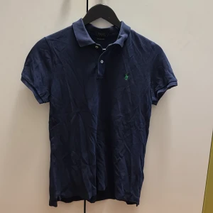 Marinblå pikétröja från Polo Ralph Lauren - Klassisk marinblå pikétröja från Polo Ralph Lauren med korta ärmar och krage. Tröjan har två knappar vid halsen och den ikoniska gröna loggan broderad på bröstet. Perfekt för en stilren och avslappnad look.