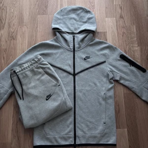 Grått Nike Tech set XS/M - Snygg grå Nike Tech set. Tröjan är i storlek S med hel dragkedja och huva. Tröjan har svarta detaljer, lång ärm och en praktisk ficka med dragkedja på ena ärmen. Perfekt för träning eller chill. Matchande byxor i storlek M medföljer, båda i mjuk bomullsblandning.