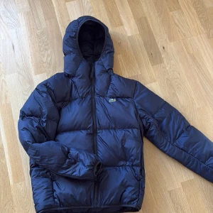 Mörkblå dunjacka/vinterjacka från Lacoste - Säljer en mörkblå dunjacka från Lacoste med huva och dragkedja. Jackan har det klassiska krokodilmärket på bröstet och är tillverkad av återvunnen polyester med fyllning av ankdun. Vattenavvisande och vindtät, perfekt för kalla dagar. Snygg, puffig look och långärmad. Säljer då den är för liten för mig.