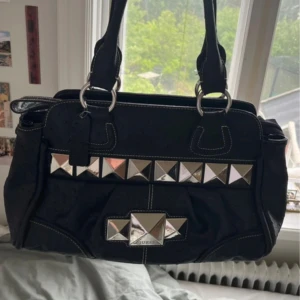 Svart handväska med nitar från Guess - Cool svart handväska från Guess med stora silvriga nitar framtill och vita kontrastsömmar. Väskan har dubbla handtag, flera fack och ett mönstrat innerfoder med Guess-logga. Materialet är präglat syntetläder som ger en lyxig känsla.