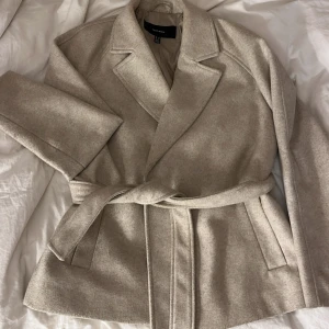 Beige kappa med bälte från Vero Moda - Snygg beige kappa från Vero Moda i klassisk modell med bred krage och knytskärp i midjan. Jackan har långa ärmar . Den är lite nopprig men det går och lösa annars inga problem