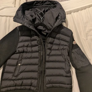 Svart cardigan från Moncler - Snygg svart pufferjacka från Moncler med stickade ärmar och vadderad kropp. Jackan har huva, dragkedja framtill, fickor med knapp och dragkedja samt Moncler-logga på ärmen. Perfekt för kyliga dagar och ger en riktigt clean look.