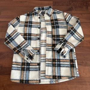 Rutig overshirt från H&M  - Overshirt från H&M i mjukt material med rutigt mönster i vitt, blått, svart och orange. Skjortan har knappar framtill, klassisk krage och långa ärmar med manschett. Perfekt för hösten.