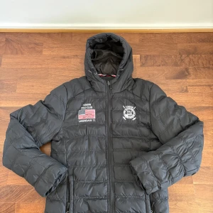 Marinblå pufferjacka från Vinson Polo Club - Säljer en svart pufferjacka från Vinson Polo Club med luva och dragkedja. Jackan har broderade detaljer på bröstet, inklusive logga och amerikansk flagga. Perfekt för kyliga dagar.