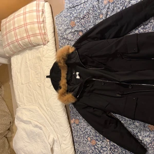 Svart parkas med fuskpäls från Sandro - Svart parkas från Sandro Paris med huva och snygg fuskpäls i beige runt kragen. Jackan har dragkedja och knappar framtill, flera fickor och en kedjedetalj vid nacken. Perfekt för kalla dagar och riktigt stilren look.