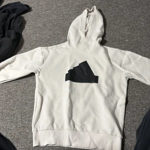 Vit hoodie från Adidas med svart logga - Säljer en stilren vit hoodie från Adidas med stor svart logga på bröstet. Tröjan har huva, långa ärmar och är tillverkad i mjuk bomullsmix. Ni står för frakten!
