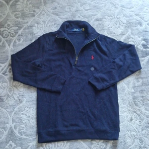 Polo Ralph Lauren half zip - Säljer en marinblå stickad tröja från Polo Ralph Lauren med halv dragkedja och klassisk röd broderad logga på bröstet. Tröjan har hög krage och långa ärmar, perfekt för kyligare dagar. Materialet är mjukt och bekvämt.