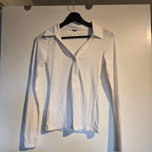 Vit långärmad polotopp med knappar - Klassisk vit långärmad polotopp med krage och tre knappar framtill. Toppen har en slim passform och är tillverkad i mjuk bomull, perfekt för en clean och stilren look. Från Vero Moda 
