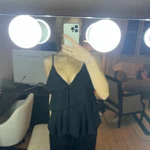 Svart volangig linnetopp med smala band - Säljer en svart linnetopp med flera lager volanger och smala axelband. Toppen har djup v-ringning och en flowy passform som ger en snygg siluett. Perfekt för dig som gillar en stilren men ändå lekfull look.