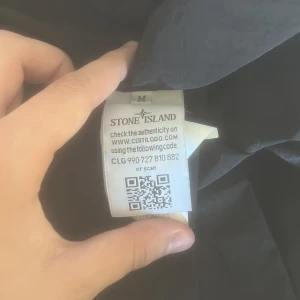 Svart overshirt från Stone Island M - Svart overshirt från Stone Island med klassisk logga på ärmen. Jackan har knäppning framtill, två bröstfickor med lock och är tillverkad i ett lätt syntetmaterial. Perfekt för dig som vill ha en clean och stilren look.