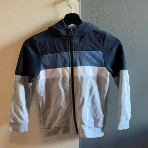 Färgblockad hoodie från BOSS - Snygg hoodie från BOSS med färgblock i mörkblått, vitt och ljusgrått. Tröjan har dragkedja framtill, huva och långa ärmar. Perfekt för dig som gillar stilren streetwear med en sportig touch.