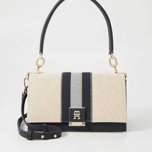 Randig axelväska från Tommy Hilfiger - Snygg axelväska från Tommy Hilfiger i beige och marinblått med breda ränder och guldfärgade metalldetaljer. Väskan har både kort och lång axelrem, praktiskt fack med dragkedja och TH-logga framtill. Tillverkad i skinn och textil för en stilren look. Använd en gång dustbag och allt ingår.