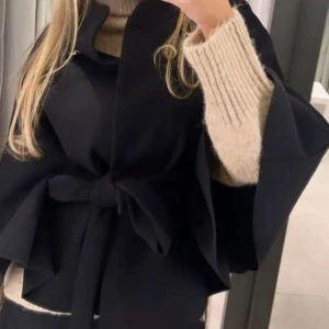 Poncho kappa från missäy - Stilren svart kappa med knytbälte i midjan och vida ärmar. Hög krage med dekorativ knapp upptill ger en elegant touch. Perfekt för dig som vill ha en trendig och sofistikerad look. Passformen är loose och materialet ser ut att vara ullblandning.