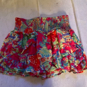 Färgglad blommig kortkjol med volang - Superfin kortkjol med färgglatt blommigt mönster i rött, grönt, blått och gult. Kjolen har bred smock i midjan och volangig nederdel som ger en luftig och lekfull känsla. Perfekt för varma dagar och somriga outfits.