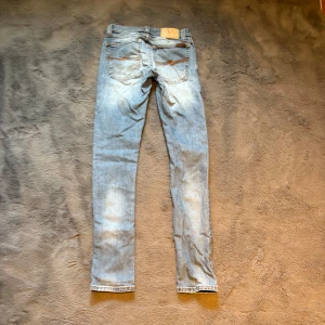 Ljusblå skinny fit jeans från Nudie Jeans - Säljer ett par ljusblå skinny fit jeans från Nudie Jeans med klassisk femficksdesign och snygga kontrastsömmar. Jeansen har en smal passform och är tillverkade i mjukt denimtyg med orange broderi på bakfickorna. Perfekt för dig som gillar skinny/slim jenas. Storlek: W:25, L:32