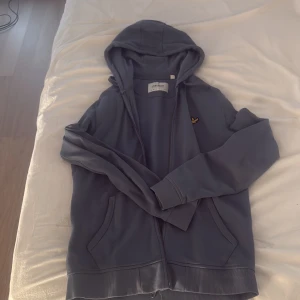 Blå hoodie från Lyle & Scott - Säljer en blå hoodie från Lyle & Scott i storlek S. Tröjan har dragkedja, huva med snörning och två fickor framtill. Klassisk gul logga på bröstet. Tillverkad i mjuk bomull, perfekt för chill dagar eller när du vill vara bekväm.