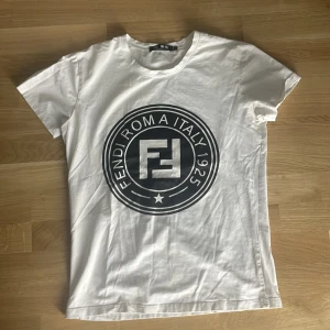 Vit Fendi t-shirt med stor logga - Vit t-shirt från Fendi med stor svart logga och texten 'Fendi Roma Italy 1925' på bröstet. Klassisk rund halsringning och korta ärmar. Tillverkad i mjuk bomull för en skön känsla. Perfekt för dig som gillar stilrena och trendiga plagg.
