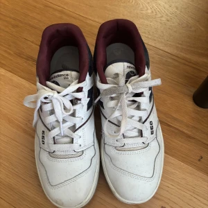 New Balance 550 vita sneakers - Säljer ett par New Balance 550 sneakers i vitt med vinröda och mörkblå detaljer. Skorna har klassisk snörning, rund tå och platt sula. Ovandelen är i skinn och textil, med tydlig 550-logga på sidan och New Balance-märke på häl och plös.