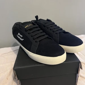 Svarta Saint Laurent sneakers - Svarta sneakers från Saint Laurent med vit broderad logga på sidan och guldfärgad text på plösen. Skorna har svart snörning, vit platt sula och är tillverkade i canvas och skinn. 