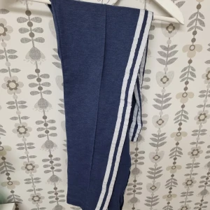 Blå mjukisbyxor adidas  - Blå mjukisbyxor från adidas med två vita ränder längs sidorna. Perfekta för en avslappnad stil och sköna att ha på sig. Byxorna har en sportig look och är gjorda i mjukt material.