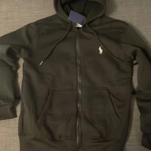 Svart hoodie - Svart hoodie med dragkedja, huva och snörning. Slim fit-modell i mjukt material med klassisk logga broderad på bröstet. Perfekt för en chill och stilren look. Fickor framtill för extra funktionalitet.