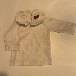 Vit långärmad tröja med krage från Mini - Säljer en söt vit långärmad tröja från Mini i mjuk bomull. Tröjan har en klassisk krage och knapp i nacken för enkel påklädning. Perfekt för dig som gillar stilrena och enkla plagg till de minsta.