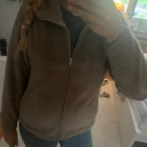 Beige teddyjacka med dragkedja - Mysig beige teddyjacka med hel dragkedja framtill och hög krage. Jackan har en loose passform och är tillverkad i fluffigt fleece-material som håller dig varm under kyliga dagar. Perfekt för dig som gillar en avslappnad och trendig stil.