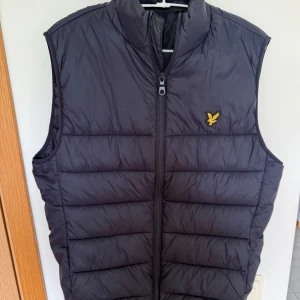 Blå dunväst från Lyle & Scott - Säljer en blå dunväst från Lyle & Scott med gul broderad logga på bröstet. Västen har quiltad design, hög krage och dragkedja framtill. Perfekt att slänga över en hoodie eller tröja för extra värme under kyliga dagar.