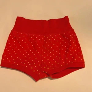 Röda shorts med hjärtan från H&M - Söta röda shorts från H&M med små gula hjärtan över hela tyget. De har en bred, mjuk resår i midjan och är tillverkade i bomull för en skön känsla. Perfekta för varma dagar när du vill ha något bekvämt och gulligt.