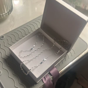 Silvrigt tvinnat halsband i presentask - Säljer ett stilrent halsband i silverfärgad metall med tvinnad kedja. Halsbandet har en snygg glansig finish och levereras i en vit presentask med lila sidenband. Perfekt accessoar för att lyfta din look.