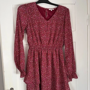 Blommig vinröd klänning med volang - Supersöt vinröd kortklänning med småblommigt mönster i rosa och vitt. Klänningen har v-ringning, långa ärmar med lätt volang vid ärmslut och markerad midja. Funkar för alla årstider. Aldrig använd. 