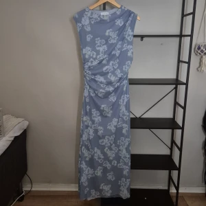 Blommig långklänning från & Other Stories - Ljusblå långklänning från & Other Stories med vitt blommönster. Klänningen har ärmlös design, draperad detalj i midjan och rund halsringning. Perfekt för sommarens alla tillfällen. Materialet känns mjukt och svalt mot huden.