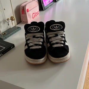 Adidas Campus svarta sneakers mocka - Svarta Adidas Campus sneakers i mocka med vita ränder och vit sula. Klassisk retrostil med tydlig Adidas-logga på plösen och CAMPUS-tryck på sidan. Skorna har snörning och en platt, gummisula som ger en clean look. Perfekta för dig som gillar streetwear och tidlös design. Lite smutsiga framtill men inget som inte går bort🤗