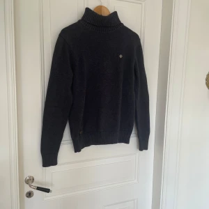 Mörkgrå stickad Morris tröja  med emblem - Mysig mörkgrå stickad morriströja med ribbad krage och ärmslut. På bröstet finns ett litet broderat emblem i vitt. Perfekt för kyliga dagar!