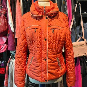 Orange quiltad jacka från Flash Woman - Snygg orange quiltad jacka från Flash Woman med hög krage, dragkedja och knappar framtill. Jackan har flera fickor med dragkedja och coola detaljer som mönstrad sömnad och dragkedjor vid ärmsluten. Perfekt för dig som vill sticka ut i höst och vinter!